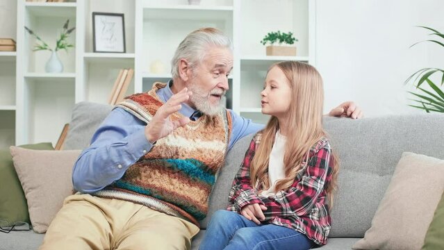 Clever caucasian kid telling beautiful poem learned by heart to loving grandfather. Kind pensioner listening attentively and giving chef's kiss. Affectionate child and granddad spending time indoors.