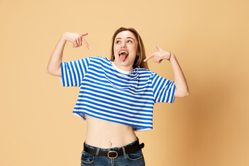 Funny, grimacing face. Portrait of emotive young girl in striped t-shirt posign with tongue sticking out against yellow studio background. Winner. Concept of youth, human emotions, facial expression