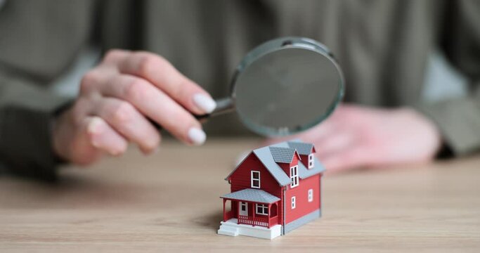 Woman looks at red house model through magnifier glass on wooden desk. Inspection of property building before purchase. Insurance concept slow motion