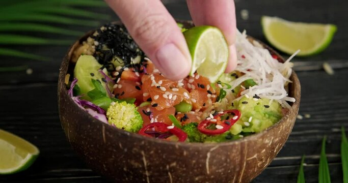 Close up poke bowl video, person preparing and serving poke dish with salmon cubes, edamame beans, cucumbers, squeezing lime juice over the dish in coconut bowl, high quality video, 4k footage