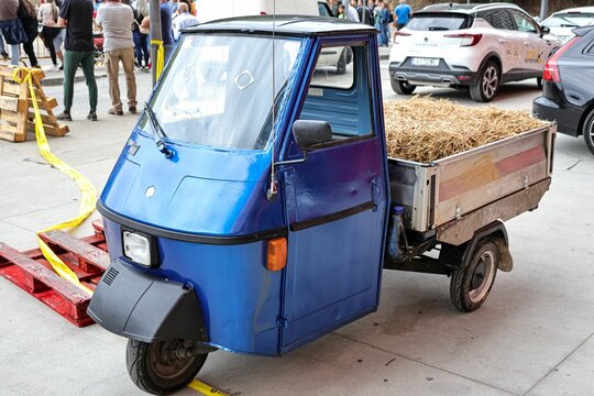 Blue vintage Piaggio Ape tricycle with a sidecar fill with hay, parked on the street