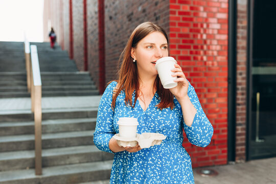 Cheerful young woman wearing blue dress walking outdoors, happy girl holding two takeaway coffee cups on city background. Urban lifestyle concept. Coffee to go. Enjoy moment, take a break.