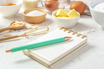 Notepad and pen for recipe with baking or cake ingredients in bowls, butter, nuts, eggs, sugar on...