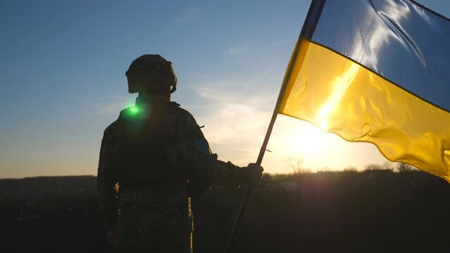 Soldier of ukrainian army holding waving flag of Ukraine against background sunset. Man in military uniform and helmet lifted up flag. Victory against russian aggression. Invasion resistance concept