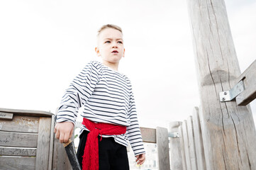 A little boy dressed as a pirate stands on the deck of a wooden ship in the playground