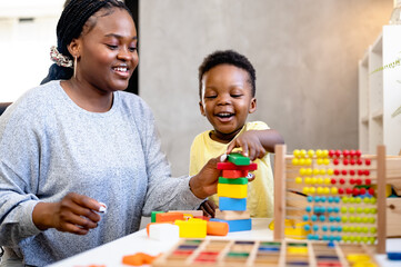 A cute little African child plays with colorful didactic educational toys. His proud mother supports him. Kindergarten teacher with child.