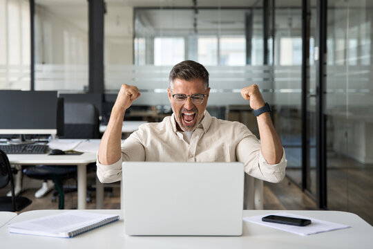 Middle-aged Latin or indian businessman screaming proud cheering with excitement , man read great news on smartphone celebrating victory and success sitting front of laptop computer working at office. - Powered by Adobe