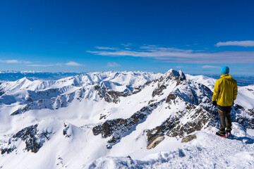 Naklejka na ściany i meble A Tourist standing on a mountain top above the clouds and looking at the view. Direct sunlight. Clear blue sky. Winter. Mountaineering. High Tatras mountain in Poland and Slovakia