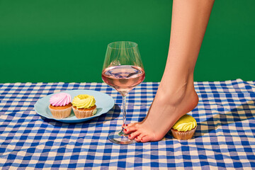 Female bare feet and delicious muffins, holding rose wine glass against green background. Party snacks, celebration. Concept of pop art photography, creative vision, imagination. Minimal art