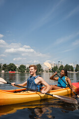 Fototapeta na wymiar positive and active multiethnic couple in life vests spending recreation weekend by sailing in sportive kayak on city river under blue sky with white clouds