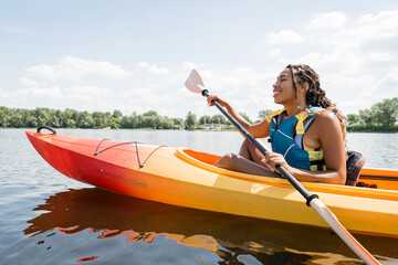 side view of carefree and active african american woman in life vest holding paddle while sailing in kayak on lake with green picturesque shore in summer - obrazy, fototapety, plakaty