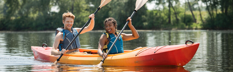smiling and active multiethnic friends in life vests paddling in sportive kayak during water recreation weekend on picturesque river on summer day, banner - obrazy, fototapety, plakaty