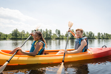 happy and charming african american woman with young and redhead man holding paddles and sailing in sportive kayak on scenic lake on active summer weekend