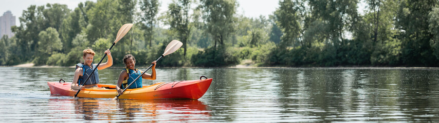 Fototapeta na wymiar cheerful african american woman and young sportive man in life vests spending time on picturesque river while sailing in kayak along green bank, banner