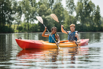 charming african american woman with young and redhead man in life vests smiling while paddling in sportive kayak on lake with green trees on shore in summer - obrazy, fototapety, plakaty