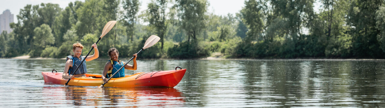 cheerful african american woman and young sportive man in life vests spending time on picturesque river while sailing in kayak along green bank, banner