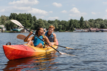 overjoyed african american woman and young redhead man in life vests spending summer weekend by sailing in kayak with paddles on lake with green scenery shore - obrazy, fototapety, plakaty