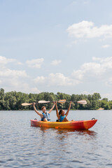 couple of cheerful interracial friends in life vests holding paddles in raised hands while sitting in kayak on picturesque lake under blue cloudy sky with green riverside on background