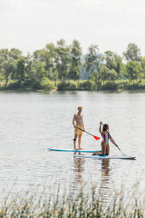 Fototapeta na wymiar redhead and sportive man paddleboarding along green riverside near african american woman in colorful swimsuit during summer recreation on summer weekend
