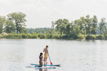Fototapeta na wymiar active multiethnic couple in colorful swimwear sailing on sup boards with paddles while spending summer vacation day on river with green trees on scenic bank