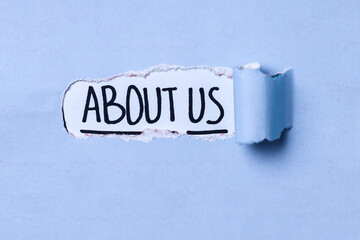 Text About Us appearing behind torn blue paper. About Us words written under ripped and torn paper