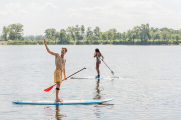 cheerful redhead man looking away and pointing with hand near sportive african american woman in colorful swimsuit sailing on sup board on summer day with picturesque riverside on background