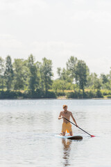 Fototapeta na wymiar young and active man in yellow swim shorts holding paddle and kneeling on sup board while sailing on scenic river with green trees on bank with picturesque riverside on background