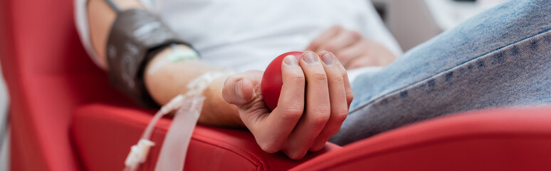 partial view of young volunteer sitting on comfortable ergonomic chair and squeezing rubber ball during medical procedure in blood donation center, banner - obrazy, fototapety, plakaty