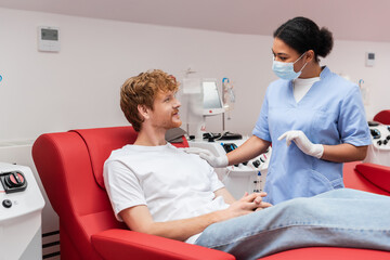multiracial nurse in medical mask and latex gloves touching shoulder and talking to redhead blood donor on medical chair near transfusion machine in clinic