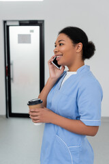 cheerful multiracial healthcare worker in blue uniform holding paper cup with takeaway coffee and...