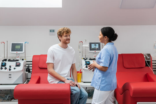 multiracial nurse in uniform talking to positive redhead volunteer sitting on medical chair near transfusion machines and plastic cup in blood donation center