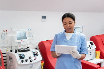 multiracial healthcare worker in blue uniform and latex gloves using digital tablet near medical...