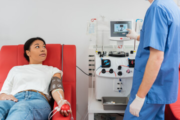 doctor in blue uniform and latex gloves operating transfusion machine near multiracial woman...