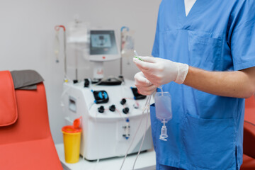 Fototapeta na wymiar cropped view of doctor in blue uniform and sterile latex gloves holding blood transfusion set near blurred automated equipment and plastic cup in laboratory