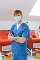 Fototapeta na wymiar young redhead doctor in medical mask and blue uniform standing with folded arms and looking at camera near medical chairs with ergonomic design and trash buckets in blood donation center