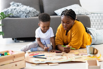 Cheerful African-American mother and cute toddler son playing with colored pencils, drawing on a big piece of paper, and sitting on the floor. Mother-son fun bonding time. Daycare creative activities.