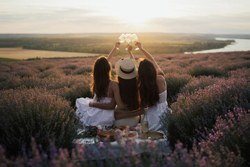 Group of girl friends with wine glasses chilling on picnic blanket at summer day. They enjoying amazing sunset. Copy space.	