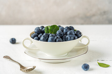 Fresh blueberries in a white teacup on the white wooden table with grey concrete background, closeup, free space for text