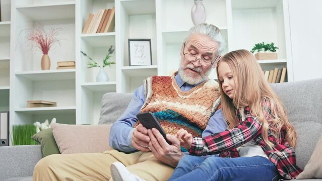Sweet little girl playing on modern cellphone while having rest on comfortable sofa with kind grandfather indoors. Long-haired child showing pictures and video kept in gallery to senior man.
