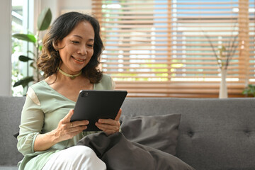 Domestic life concept with middle aged Asian woman looking happy using digital tablet while sitting on sofa.