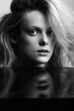 Beauty, fashion and make-up concept. Beautiful and young blonde woman with big wavy hair close-up studio portrait. Model face reflection on black mirror. Sensual mood. Black and white image