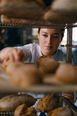 attractive female baker between shelves looking and checking freshly baked bread very carefully in...