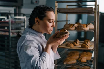 Tuinposter attractive female baker takes freshly baked fragrant bread from the shelf sniffs it checks its quality bakery professional kitchen baking © Guys Who Shoot