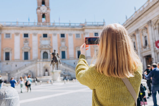 Blonde Woman with smartphone is walking on a sunny day. Capitol in Rome, Piazza del Campidoglio in Capitoline Hill, Rome, Italy. Concept of traveling famous landmarks.