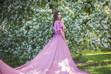 Fototapeta premium A young beautiful brunette girl stands near a branchy spring apple tree. A girl in a pink dress smiles between blooming apple trees. The concept of spring or summer