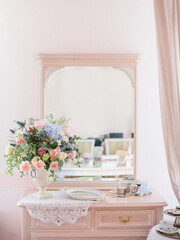 Women's pink dressing table in vintage style with a mirror. On the dressing table a bouquet of peonies, roses and hydrangeas, a metal tray, a box of pearls, perfume. Company faux french style.