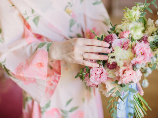 A bride in a pink robe with flowers holds a pink wedding bouquet in her hands. Wedding ring on the bride's finger.