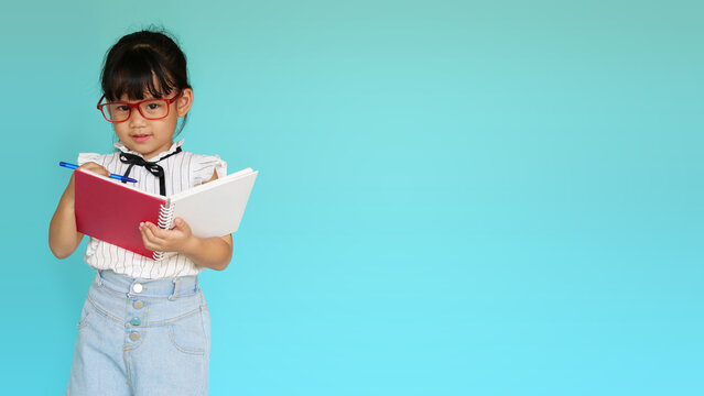 Cute student girl smiling and holding a book. Portrait of happy Asian glasses girl writing on the notebook with isolated green background. Closeup face of schoolgirl looking at camera. Education.