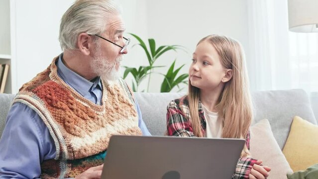Grey-haired aged man surfing internet with curious granddaughter in order to find food delivery. Loving grandpa offering online restaurant on wireless computer and little girl nodding with agreement.