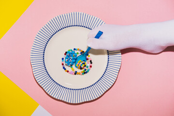 A foot in a white tabi sock holds a blue plastic spoon over colored necklace beads in the form of food over a large plate. Creative concept, top view - 606503219